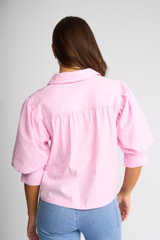 A woman viewed from the back wearing a light pink textured blouse with puff sleeves and light blue jeans, set against a plain light gray studio background.