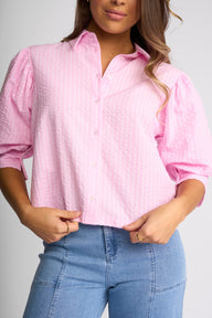 A woman wearing a light pink textured button-up blouse with puffed three-quarter sleeves and light blue jeans, shown from the nose down against a light gray studio background.