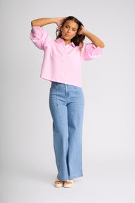 A woman modeling a light pink cropped button-up shirt with textured vertical stripes and light-wash blue wide-leg jeans, standing against a light gray studio background.