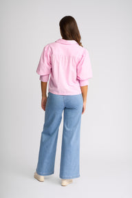 A woman shown from the back wearing a light pink short-sleeve button-up shirt and light blue wide-leg jeans, standing against a light gray studio background.