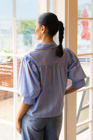 A woman with her hair in a single braid is shown from behind, wearing a light blue striped blouse with thin gold and orange vertical stripes, paired with blue jeans, standing in front of a large window.
