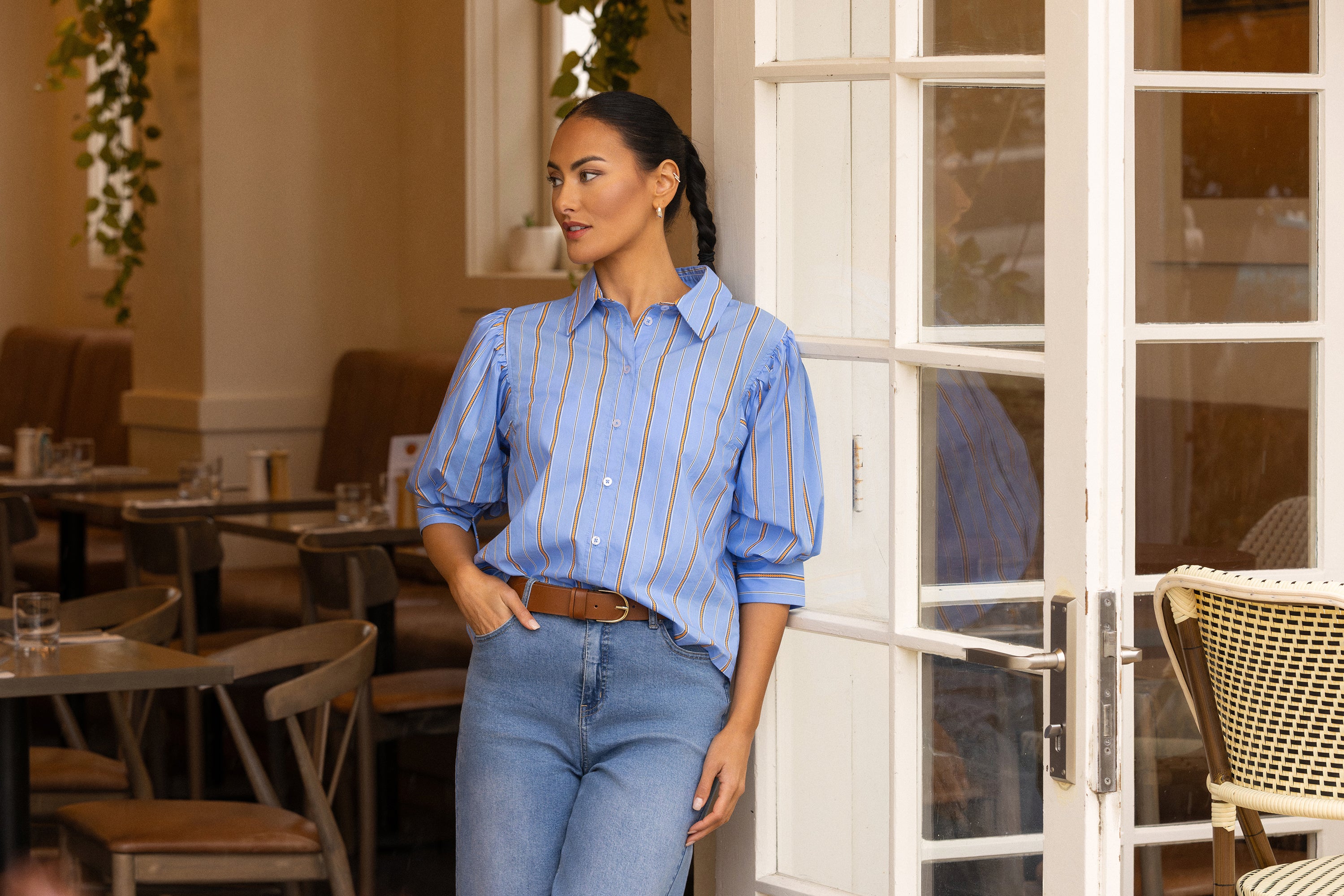 A woman stands in a cafe doorway wearing a light blue button-up shirt with thin vertical tan stripes, paired with blue jeans and a brown belt.