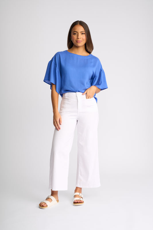 A woman modeling a bright blue short-sleeve top with voluminous flutter sleeves, paired with white wide-leg cropped pants and beige slide sandals, against a light gray studio background.