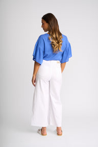 A woman shown from the back wearing a bright blue short-sleeve blouse with ruffled sleeves and white wide-leg cropped pants, standing against a light gray studio background.