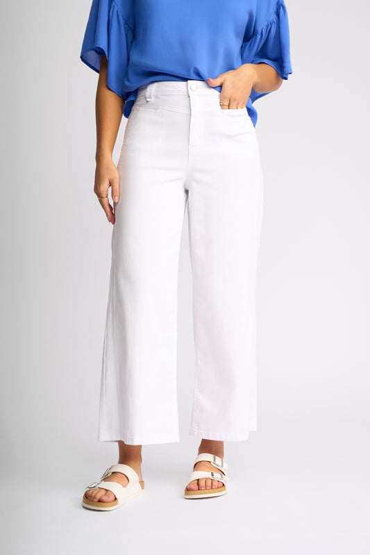 A model wearing white cropped wide-leg pants, a blue blouse, and white slide sandals, shown from the waist down against a light gray studio background.