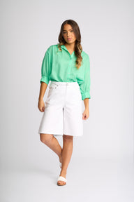 A woman modeling a mint green button-up blouse with rolled sleeves and white knee-length shorts against a light gray studio background.