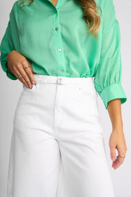 A woman wearing a mint green button-up blouse with cuffed sleeves and white high-waisted trousers, shown from the chest down against a light gray studio background.