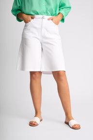A model wearing white high-waisted shorts with a matching white belt, paired with a green button-up shirt and white slide sandals, shown from the waist down on a light gray studio background.