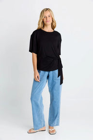 A woman modeling a black short-sleeve top with a side twist detail, paired with light blue jeans and white sandals, against a plain light gray studio background.