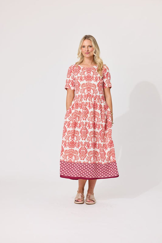 A woman modeling a short-sleeved midi dress with a red and white floral pattern and a solid magenta border, standing against a light gray studio background.