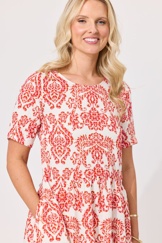 A smiling blonde woman wearing a short-sleeve dress with a red and white damask pattern, photographed against a light gray studio background.
