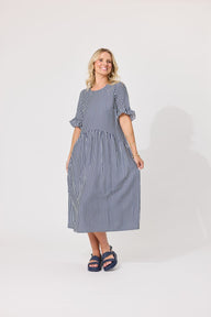 A woman modeling a blue and white vertically striped midi dress with ruffled short sleeves and side pockets, paired with navy blue sandals, against a plain light gray studio background.