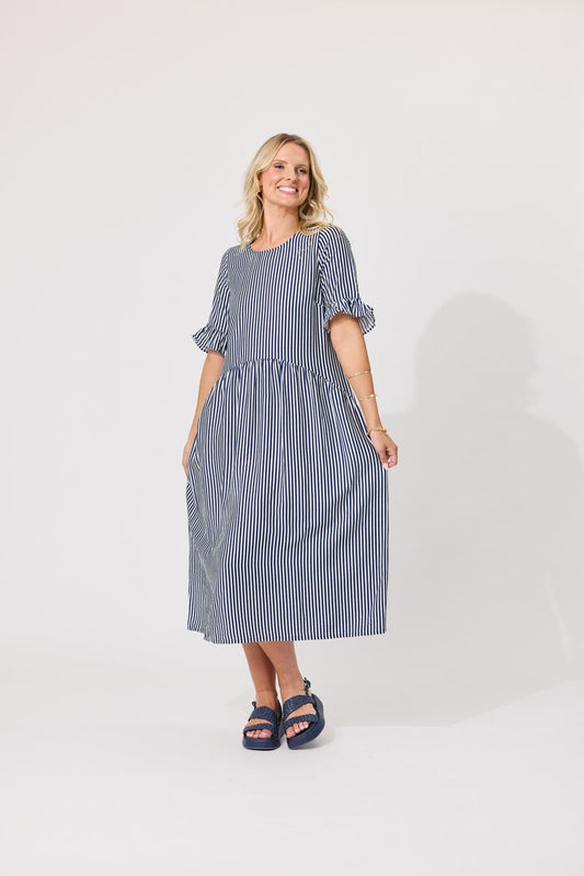 A woman modeling a blue and white vertically striped midi dress with ruffled short sleeves and side pockets, paired with navy blue sandals, against a plain light gray studio background.