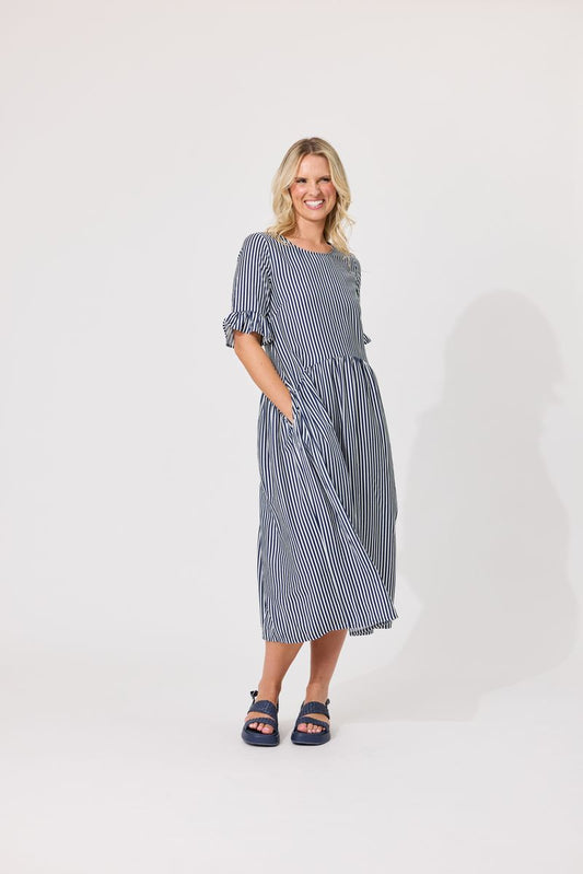 A woman modeling a blue and white vertically striped midi dress with ruffled three-quarter sleeves and side pockets, paired with dark blue slide sandals, against a plain light gray studio background.