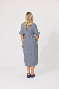 A woman viewed from the back wearing a blue and white vertically striped midi dress with ruffled short sleeves and dark navy sandals, standing against a plain white studio background.