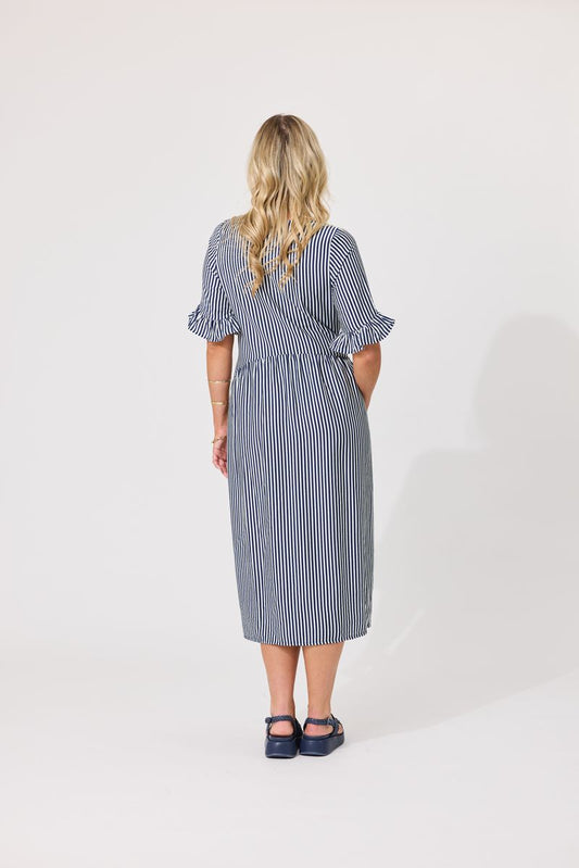 A woman viewed from the back wearing a blue and white vertically striped midi dress with ruffled short sleeves and dark navy sandals, standing against a plain white studio background.