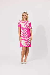A woman modeling a short-sleeve collared dress with a bold pink and off-white palm leaf pattern, standing against a plain light gray studio background.
