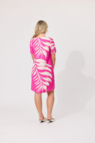 A woman viewed from behind wearing a short-sleeve pink dress with a large cream tropical leaf print, standing against a light gray studio background.