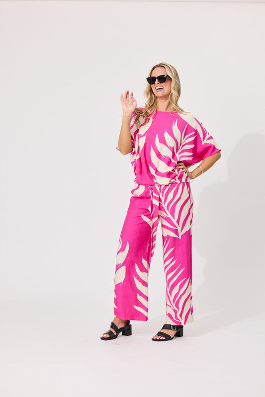 A woman modeling a bright pink two-piece set with a white palm leaf pattern, consisting of a short-sleeve top and matching wide-leg pants, styled with black sunglasses and black sandals against a plain white studio background.