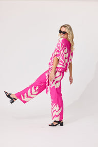 A woman modeling a bright pink and white tropical leaf print two-piece set consisting of a short-sleeve tunic top and matching wide-leg pants, styled with black sandals against a plain white studio background.
