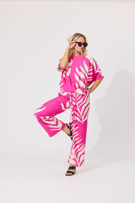 Bright pink wide-leg pants with a bold cream palm leaf print, modeled with black strappy sandals against a plain white studio background.