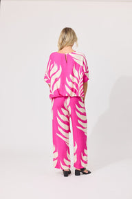 A woman viewed from behind wearing a bright pink and white tropical leaf print jumpsuit, paired with black sandals, against a plain white studio background.