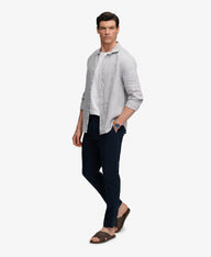 A man modeling a light gray linen button-up shirt over a white t-shirt, paired with dark navy pants and brown sandals, against a plain white background.