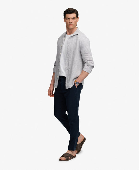 A man modeling a light gray linen button-up shirt over a white t-shirt, paired with dark navy pants and brown sandals, against a plain white background.