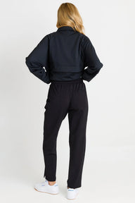A woman shown from the back wearing a black cropped jacket and black pants, paired with white sneakers, against a plain light gray studio background.