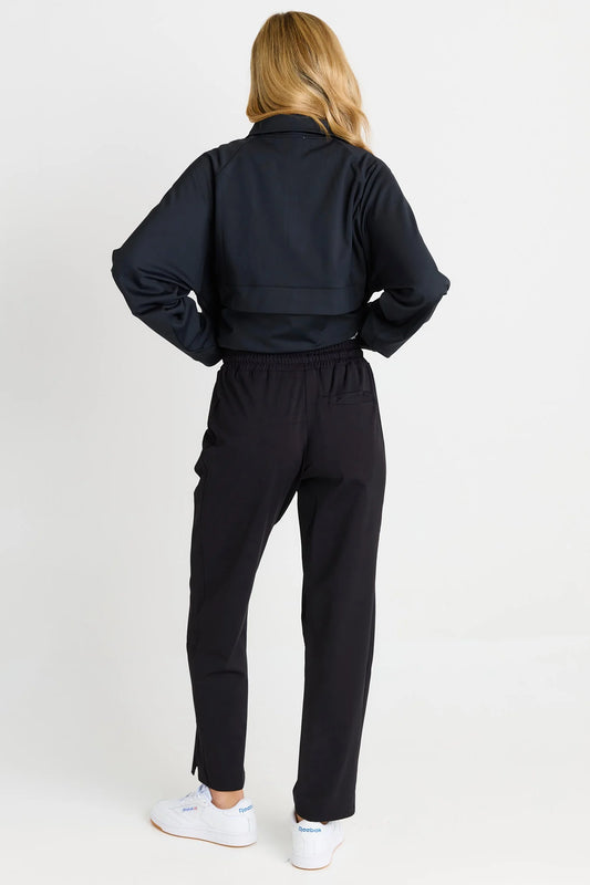 A woman shown from the back wearing a black cropped jacket and black pants, paired with white sneakers, against a plain light gray studio background.