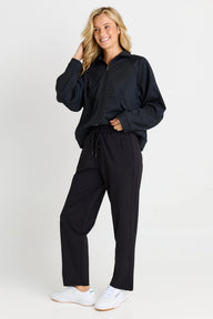 A woman modeling a black zip-up jacket and matching black drawstring sweatpants with white sneakers, photographed against a clean light gray studio background.