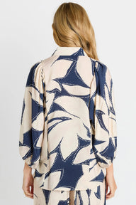 A woman viewed from the back wearing a loose long-sleeve blouse with a large abstract floral or leaf-like print in navy blue and warm cream, paired with matching bottoms against a light gray studio background.