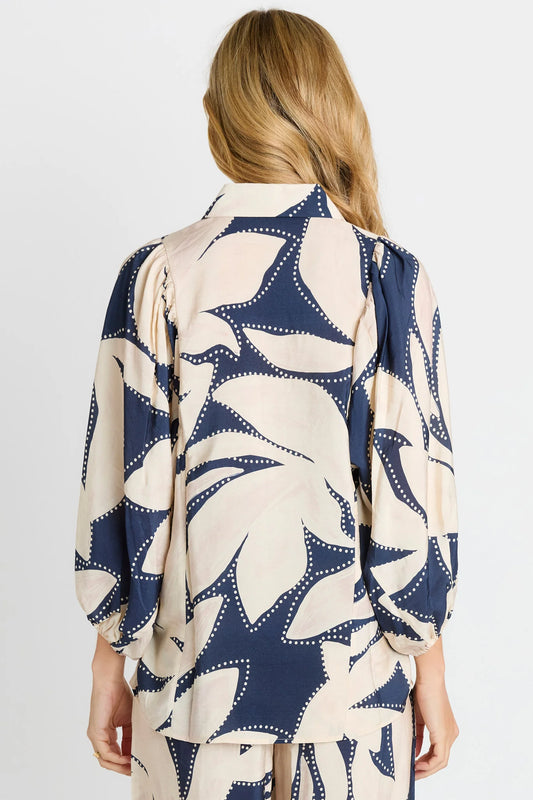 A woman viewed from the back wearing a loose long-sleeve blouse with a large abstract floral or leaf-like print in navy blue and warm cream, paired with matching bottoms against a light gray studio background.