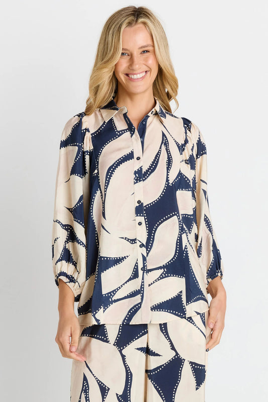 A woman modeling a matching two-piece lounge set featuring a long-sleeve button-up shirt and pants in a navy blue and cream abstract pattern, set against a plain light gray studio background.
