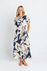 A woman modeling a navy blue and cream tropical leaf print midi dress with short sleeves, a button-up collar, and a tie waist, paired with tan sandals against a plain white studio background.
