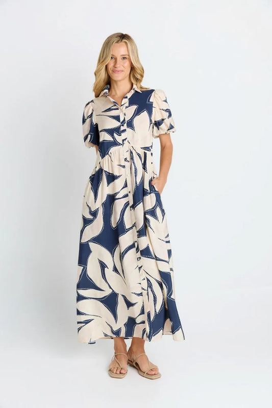 A woman modeling a navy blue and cream tropical leaf print midi dress with short sleeves, a button-up collar, and a tie waist, paired with tan sandals against a plain white studio background.