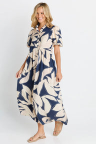 A woman modeling a navy blue and cream leaf-patterned midi dress with short puff sleeves and a button-up collar, paired with beige flat sandals, against a light gray studio background.