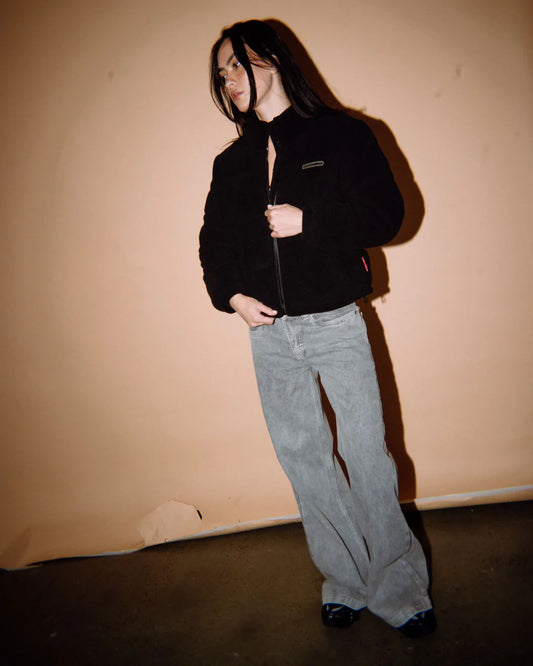 Person wearing a black jacket and gray pants against a beige wall.
