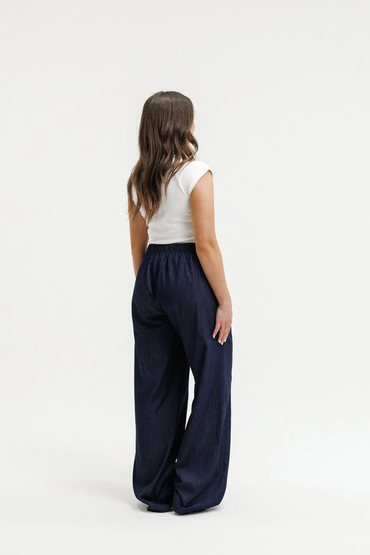 A woman shown from the back wearing a white sleeveless top and dark navy wide-leg trousers, standing against a plain white studio background.