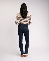 A woman viewed from behind wearing dark blue skinny jeans with contrast stitching and a beige and black striped long-sleeve top, standing against a light gray studio background.