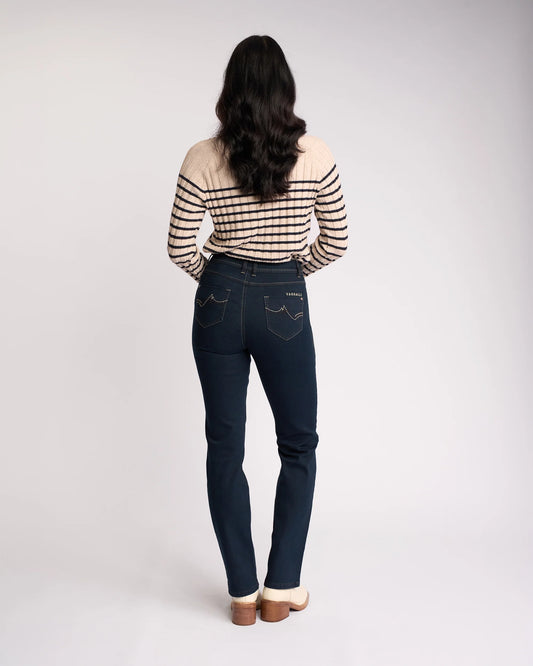 A woman viewed from behind wearing dark blue skinny jeans with contrast stitching and a beige and black striped long-sleeve top, standing against a light gray studio background.