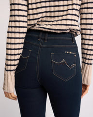 Rear view of a person wearing dark blue Vassalli skinny jeans with decorative contrast stitching and a striped beige and navy knit top, against a light gray studio background.