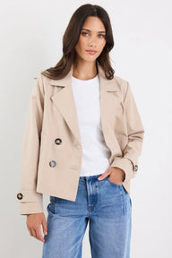 Woman wearing a beige jacket, white shirt, and blue jeans on a plain background