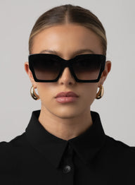 Woman wearing black sunglasses and gold hoop earrings against a gray background