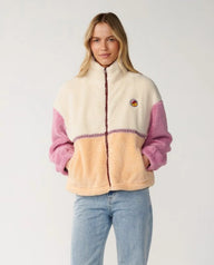 A woman modeling a sherpa fleece zip-up jacket with cream, peach, and pink color blocking, paired with light blue jeans against a plain light gray background.