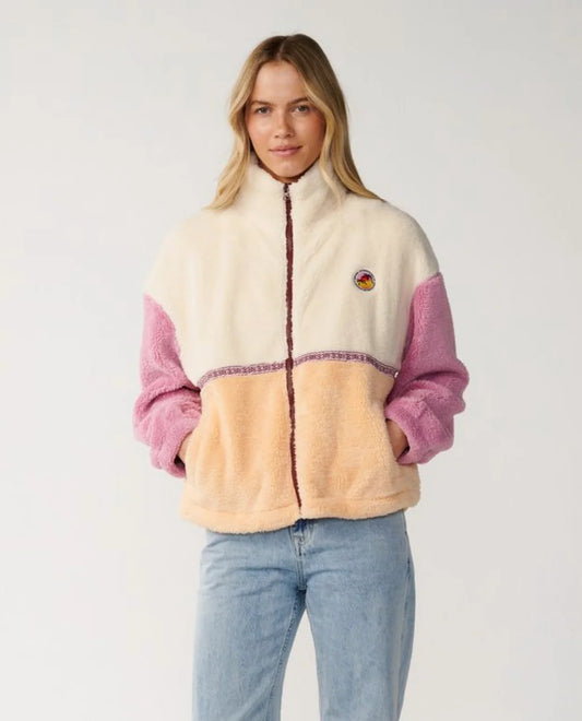 A woman modeling a sherpa fleece zip-up jacket with cream, peach, and pink color blocking, paired with light blue jeans against a plain light gray background.