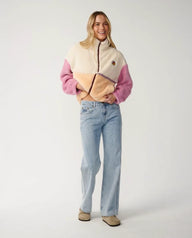 A woman modeling a color-block fleece jacket with cream, pink, and tan panels, paired with light-wash blue jeans and beige shoes, against a plain light gray studio background.