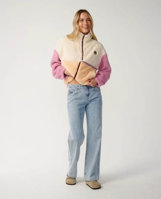 A woman modeling a color-block fleece jacket with cream, pink, and tan panels, paired with light-wash blue jeans and beige shoes, against a plain light gray studio background.