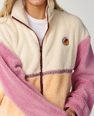 A close-up of a person wearing a sherpa fleece zip-up jacket with cream, pink, and tan color-blocked panels, a dark brown zipper, and a circular embroidered logo on the chest.