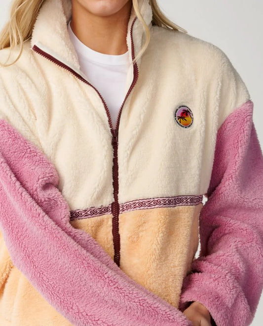 A close-up of a person wearing a sherpa fleece zip-up jacket with cream, pink, and tan color-blocked panels, a dark brown zipper, and a circular embroidered logo on the chest.